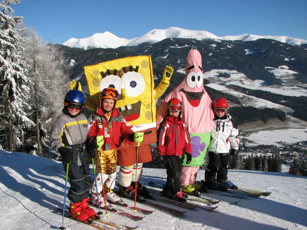 Kindergruppe im Skikurs posiert für ein Foto mit den zwei Maskottchen der Skischule im Skigebiet Gaal in der Steiermark. JUFA Hotels bietet erholsamen Familienurlaub und einen unvergesslichen Winterurlaub.
