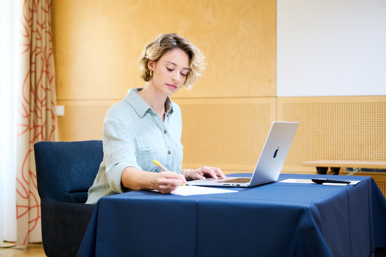 Ihr seht eine konzentrierte Frau, die etwas in einem Seminarraum ausfüllt im JUFA Hotel Wien City****. Sie sitzt auf einen dunkelblauen Sessel und vor ihr ein Apple Laptop auf einem Tisch mit einem dunkelblauen Tischtuch. Der Ort für eure erfolgreichen Seminare, Workshops und Tagungen.