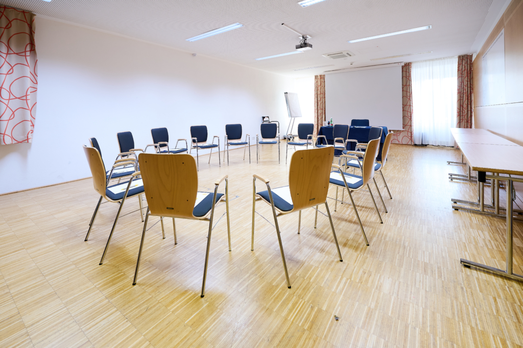 Ihr seht den Seminarraum Menuett im JUFA Hotel Wien City****. Die Bestuhlung ist in einem Sesselkreis aufgebaut. Im Hintergrund befindet sich ein Beamer, eine Leinwand und ein Flipchart. Der Ort für eure erfolgreichen Seminare, Workshops und Tagungen.